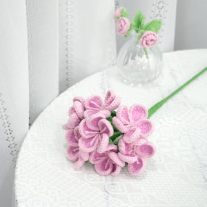 May include: A bouquet of pink crocheted flowers with green stems. The flowers are arranged in a cluster and are sitting on a white lace tablecloth.