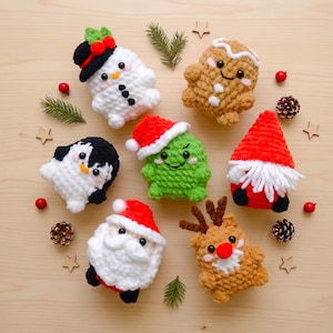 May include: A collection of seven crocheted plush ornaments including a snowman, gingerbread person, penguin, green creature in a Santa hat, gnome, Santa Claus, and reindeer, arranged with pine sprigs, pinecones, and wooden stars.