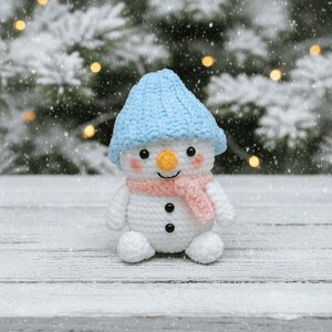 May include: A crocheted snowman toy, wearing a light blue hat and a pink scarf. The snowman has a carrot nose, black button eyes, and a smiling mouth. It sits on a snowy wooden surface, with a blurred background of a snowy evergreen tree.