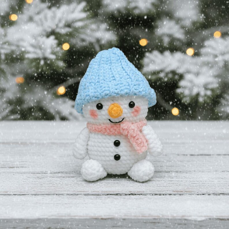 May include: A crocheted snowman toy, wearing a light blue hat and a pink scarf. The snowman has a carrot nose, black button eyes, and a smiling mouth. It sits on a snowy wooden surface, with a blurred background of a snowy evergreen tree.