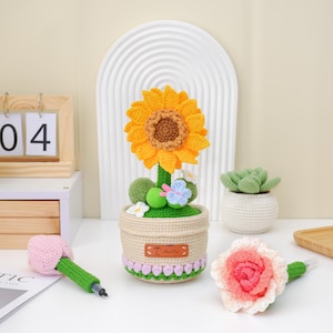 May include: A crocheted sunflower in a beige pot with green trim, a pink and green pen, and a pink and white flower. The sunflower has yellow petals and a brown center. A small green ball and a blue butterfly are also included.