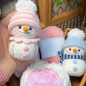 May include: A basket contains handmade crochet snowmen and balls of yarn. One snowman has a pink hat and collar, the other a blue hat and scarf. Both have carrot noses and button eyes. The yarn is pink and white.