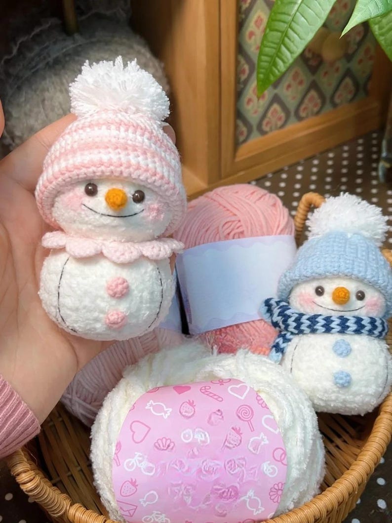 May include: A basket contains handmade crochet snowmen and balls of yarn. One snowman has a pink hat and collar, the other a blue hat and scarf. Both have carrot noses and button eyes. The yarn is pink and white.