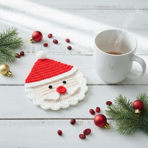 May include: A crocheted Santa Claus coaster with a red hat and white beard sits next to a white mug with a hot beverage. Red and gold Christmas ornaments and sprigs of evergreen are scattered around the coaster on a white wooden surface.