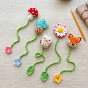 Peut inclure: Un ensemble de cinq marque-pages en crochet comprenant un champignon rouge et blanc, un cactus vert dans un pot marron, une chouette beige, une marguerite blanche et jaune, et un renard orange et blanc avec une longue tige verte et une feuille.