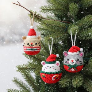 May include: Three handmade crochet cat ornaments hanging on a Christmas tree. The ornaments are beige, gray, and white with red and green accents, each wearing a Santa hat. The ornaments are suspended by twine or white string.