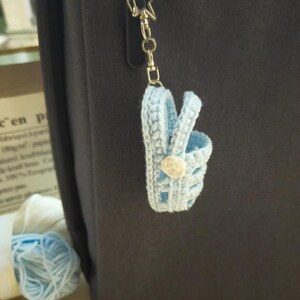 Op de afbeelding: Een lichtblauwe gehaakte miniatuur mand sleutelhanger met een klein wit bloemaccent aan de zijkant. De mand is bevestigd aan een zilverkleurige metalen stervormige sluiting en een ketting, hangend tegen een donkergrijs oppervlak.