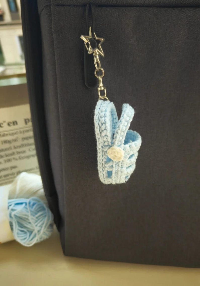 Op de afbeelding: Een lichtblauwe gehaakte miniatuur mand sleutelhanger met een klein wit bloemaccent aan de zijkant. De mand is bevestigd aan een zilverkleurige metalen stervormige sluiting en een ketting, hangend tegen een donkergrijs oppervlak.