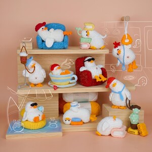 May include: A collection of small, white, crocheted duck figures in various poses and settings, including sitting on a blue chair, lounging in a teacup, wearing sunglasses on a red chair, hanging from a strap, and resting on books or a yellow float.