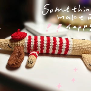 May include: A crocheted dachshund dog toy wearing a red beret and a red and white striped sweater. The dog is beige with brown paws and tail. The text "Something make me happy" is written in white cursive.