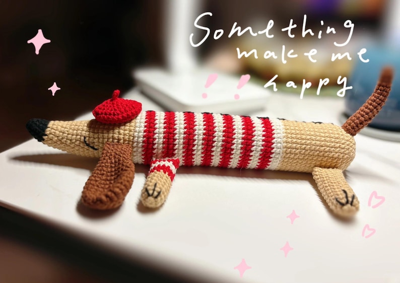 May include: A crocheted dachshund dog toy wearing a red beret and a red and white striped sweater. The dog is beige with brown paws and tail. The text "Something make me happy" is written in white cursive.