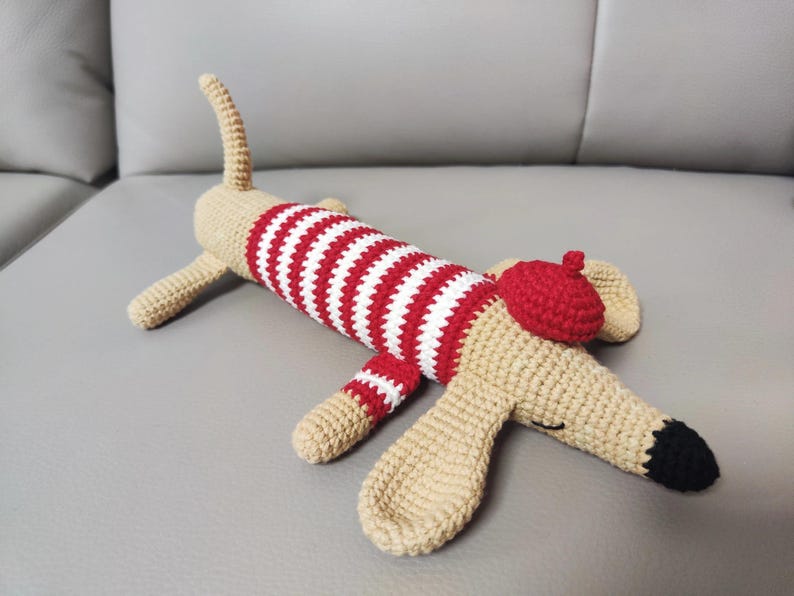 May include: A handmade crochet dog toy in beige, red, and white. The dog wears a red and white striped sweater and a red beret. The dog's nose is black, and it has a curled tail. The toy is lying on a gray surface.