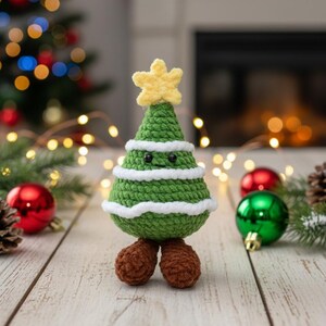 May include: A handmade green crochet Christmas tree with white accents and a yellow star on top. The tree has black eyes and brown feet. Christmas ornaments and a Christmas tree are in the background.