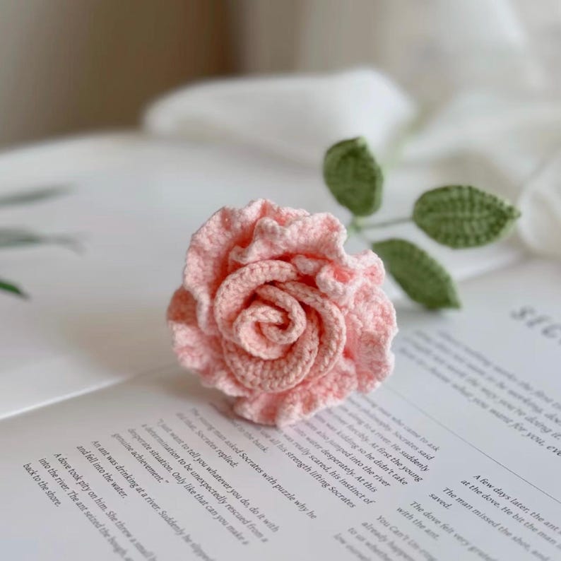 May include: A crocheted pink rose with green leaves. The rose is made with a delicate, lacy stitch and has a ruffled edge. The rose is sitting on a book with text about Socrates.