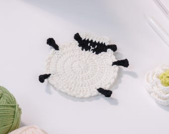 May include: A handmade, white crochet sheep coaster with black accents. The sheep has a round body, black legs, and a black and white head. The coaster is placed on a white surface, with yarn and a flower in the background.