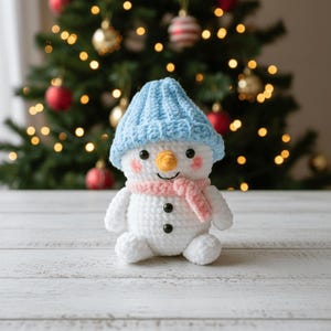 May include: A crocheted snowman in white, wearing a blue hat and a pink scarf. It has a carrot nose, black button eyes, and a smiling mouth. The snowman sits on a white wooden surface, with a Christmas tree decorated with baubles in the background.
