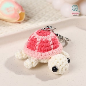 Op de afbeelding: Een gehaakte schildpad sleutelhanger met een roze en witte schild en een wit lichaam. De sleutelhanger heeft zwarte ogen en een zilveren metalen ring.