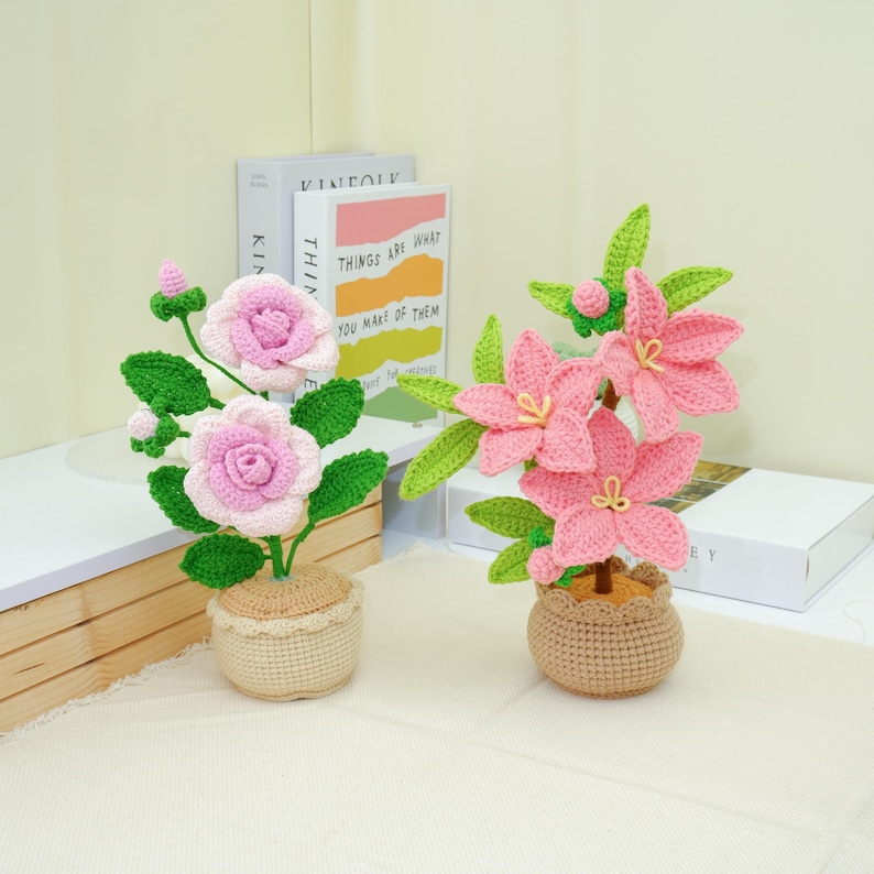 May include: Two crochet flower arrangements. One features pink roses, green leaves, and a beige pot. The other has pink lily-like flowers, green leaves, and a similar pot. Decorative items for home decor.