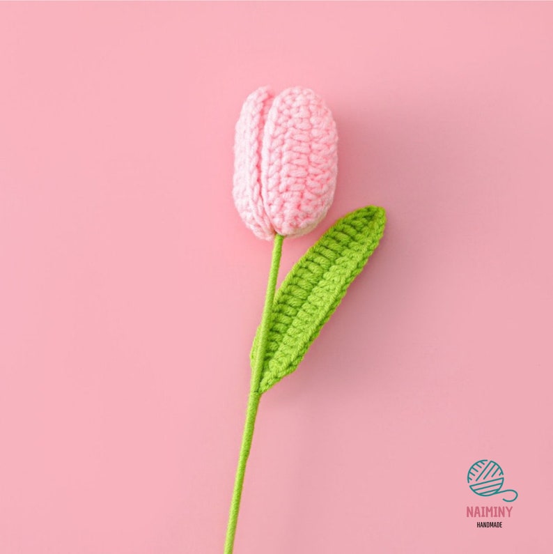 May include: A pink crocheted tulip with a green crocheted leaf and stem. The flower is on a pink background.