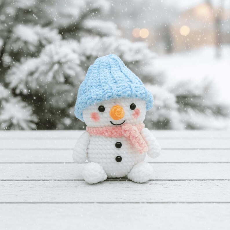 May include: A crocheted snowman wearing a light blue hat and a pink scarf. The snowman has black button eyes, an orange nose, and rosy cheeks. It is set against a snowy backdrop.