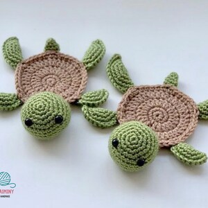 May include: Two crocheted turtle coasters in green and brown. The turtles have black eyes and are lying on a white surface.