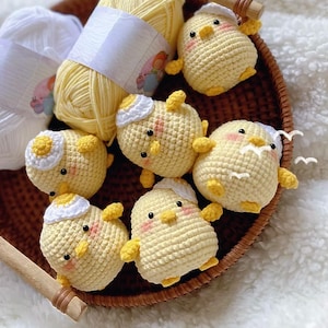 May include: A selection of handmade crochet chicks in a woven basket. The chicks are yellow with white details, small wings, and pink cheeks. Two skeins of yarn, one yellow and one white, are also in the basket. The chicks are a charming, handcrafted item.