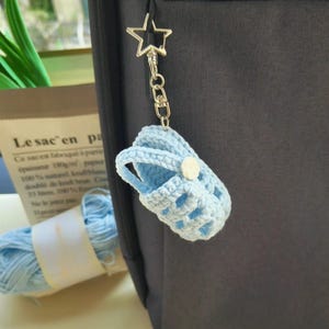 Op de afbeelding: Een lichtblauwe gehaakte babyschoen-sleutelhanger met een klein wit knoopje. De miniatuurschoen is bevestigd aan een zilverkleurige metalen stervormige sluiting en een ketting, hangend aan een donkergrijs stoffen oppervlak.
