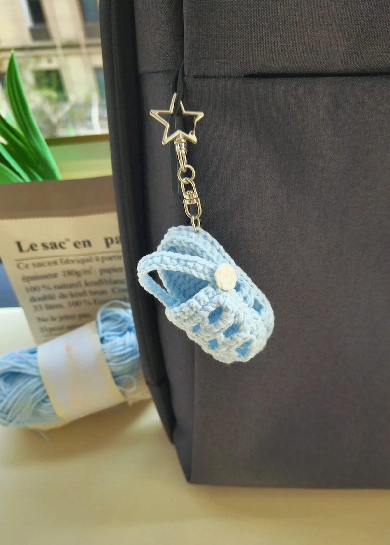 Op de afbeelding: Een lichtblauwe gehaakte babyschoen-sleutelhanger met een klein wit knoopje. De miniatuurschoen is bevestigd aan een zilverkleurige metalen stervormige sluiting en een ketting, hangend aan een donkergrijs stoffen oppervlak.