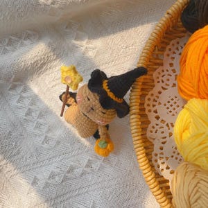 May include: A crocheted witch doll with a black hat, holding a star-topped wand and a small pumpkin. The doll is tan with black accents and a smiling face. The doll is next to a basket with yarn.