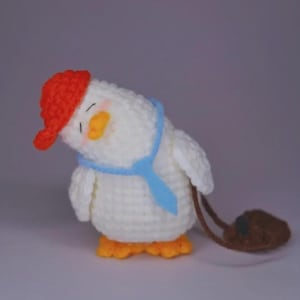 May include: A white crocheted duckling toy with a tilted head, wearing a bright red cap, a light blue necktie, and orange feet. The duckling has closed eyes with pink blush on its cheeks and carries a small brown crocheted bag on its side.