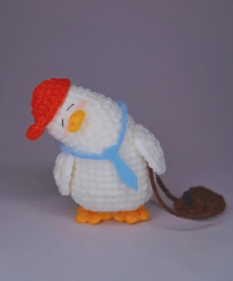 May include: A white crocheted duckling toy with a tilted head, wearing a bright red cap, a light blue necktie, and orange feet. The duckling has closed eyes with pink blush on its cheeks and carries a small brown crocheted bag on its side.