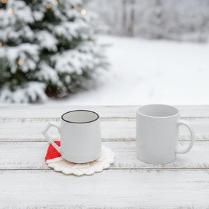 May include: Two white ceramic mugs on a white wooden surface. One mug has a black rim and sits on a red and white crocheted coaster. A snowy winter scene with a Christmas tree is in the background.