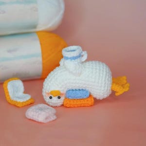 Puede incluir: Un pato blanco hecho a mano con ganchillo con un asiento azul y naranja, una pequeña taza de té y plumas de cola amarillas. Las piezas de ganchillo adicionales incluyen un asiento y una pequeña almohada rosa. Dos madejas de hilo están en el fondo.