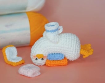 May include: A handmade white crochet duck with a blue and orange seat, a small teacup, and yellow tail feathers. Additional crochet pieces include a seat and a small pink pillow. Two yarn skeins are in the background.