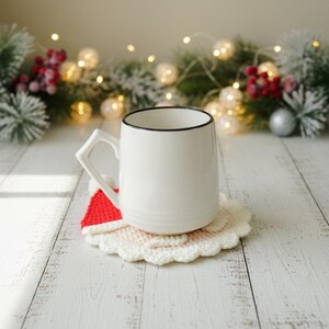 May include: A white ceramic mug with a black rim sits on a handmade, crocheted coaster. The coaster features a red and white Santa hat design. The background includes festive greenery, fairy lights, and decorative ornaments, creating a holiday theme.