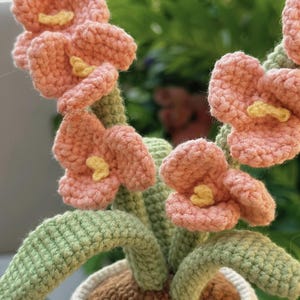 Puede incluir: Un arreglo floral de ganchillo hecho a mano en una maceta blanca. Las flores son de color melocot&oacute;n con centros amarillos, y las hojas y los tallos son verdes. La maceta tiene un interior marr&oacute;n.