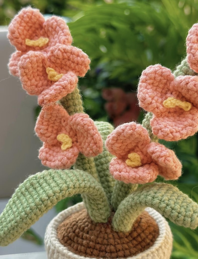 Puede incluir: Un arreglo floral de ganchillo hecho a mano en una maceta blanca. Las flores son de color melocot&oacute;n con centros amarillos, y las hojas y los tallos son verdes. La maceta tiene un interior marr&oacute;n.