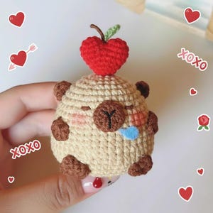 May include: A handmade crochet bear with a red heart on top. The bear is beige with brown accents and a blue tongue. The bear is holding in a hand. Decorative hearts and the word "XOXO" are in the background.