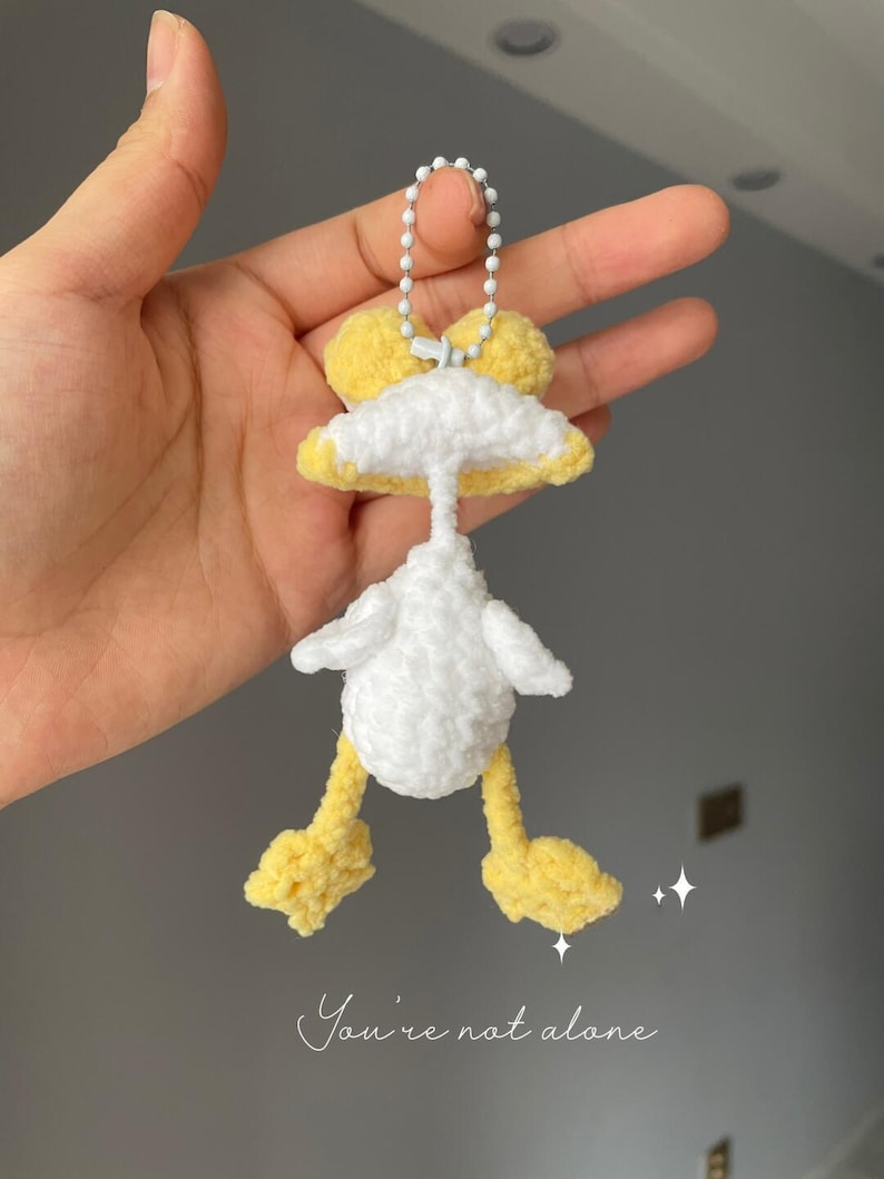 May include: A handmade crochet duck keyring in white and yellow. The duck has a white body, yellow feet and head, and a small white bow. The text "You're not alone" is written in cursive at the bottom.