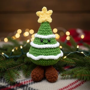 May include: A handmade green crochet Christmas tree with a yellow star on top. The tree has white trim and brown feet. The tree has a smiling face with black eyes. The tree is sitting on a plaid blanket with pine needles.