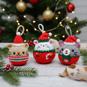 May include: Three crocheted Christmas ornaments shaped like mice, each wearing a Santa hat. The ornaments are in beige, white, and gray, with red and green accents. They have embroidered faces and are hanging in front of a Christmas tree.