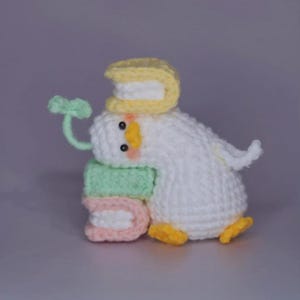 May include: A handmade crochet duck figurine with a yellow hat, yellow feet, and a green stem. The duck is white with a yellow beak and small black eyes. It is holding a stack of colorful, U-shaped objects.