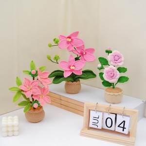 May include: Handmade crochet flower arrangements featuring pink orchids and roses in small woven pots. The flowers have green leaves and stems. A wooden calendar displays "JUL 04". A white, square, decorative candle is in the lower left corner.
