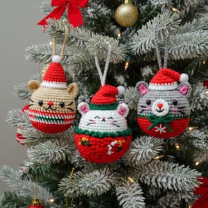 May include: Three crocheted Christmas ornaments shaped like mice, each wearing a Santa hat. The ornaments are beige, white, and gray, with red and green accents. They hang on a decorated Christmas tree.