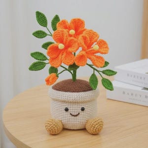May include: A crocheted potted plant featuring bright orange flowers with yellow centers and green leaves. The pot is cream-colored with a brown top, a stitched smiling face, and two small tan feet at the base, crafted from yarn in a textured style.