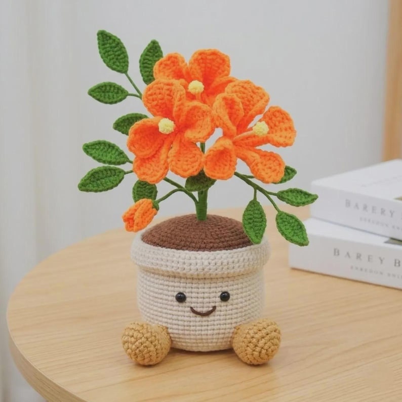 May include: A crocheted potted plant featuring bright orange flowers with yellow centers and green leaves. The pot is cream-colored with a brown top, a stitched smiling face, and two small tan feet at the base, crafted from yarn in a textured style.