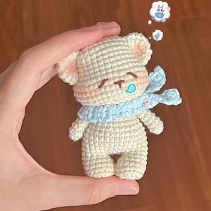 Könnte beinhalten: Ein handgehäkelter Teddybär aus cremefarbenem Garn, der einen hellblauen Schal trägt. Der Bär hat geschlossene Augen, einen blauen Tropfen und rosa Innenohren. Eine Sprechblase zeigt ein Kaninchen. Der Bär wird in einer Hand gehalten.