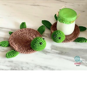 May include: Two crocheted turtle-shaped coasters in brown and green. The coasters are handmade and feature black eyes. The coasters are on a white surface.