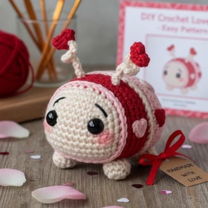 Könnte beinhalten: Eine handgefertigte Häkel-Marienkäfer-Figur mit rot-rosa gestreiftem Körper, Herzakzenten und großen schwarzen Augen. Der Marienkäfer hat Fühler mit roten Herzspitzen. Ein Etikett lautet "Handmade with Love".