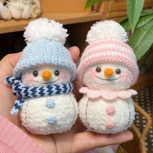 May include: Two handmade snowman ornaments. One wears a blue knit hat and scarf, the other a pink hat and collar. Both have orange carrot noses, rosy cheeks, and stitched smiles. They are made of white plush material.